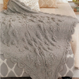 Könnte beinhalten: Eine graue gestrickte Wolldecke mit einem Blatt-Muster. Die Decke liegt auf einem Sofa mit gemusterten Kissen.