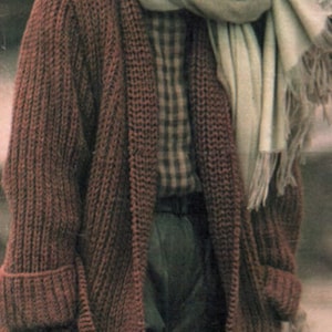 以下が含まれることがあります： ショールカラーと白いスカーフが付いた、長い茶色のニットカーディガン。カーディガンはゆったりとしたフィット感で、太い糸で作られています。