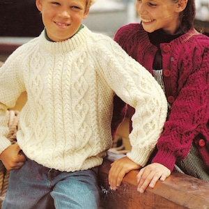 May include: Two children wearing knitted sweaters. The boy is wearing a cream colored cable knit sweater. The girl is wearing a burgundy cable knit cardigan with buttons.