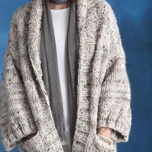 Könnte beinhalten: Ein weißer und grauer Strickcardigan mit Schalkragen und Taschen. Der Cardigan ist aus einem dicken Garn gefertigt und hat eine lockere Passform.