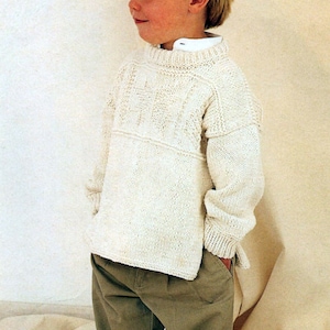 May include: A young boy wearing a white knitted sweater with a cable knit pattern and brown pants.