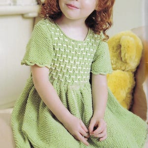 May include: A young girl wearing a green knitted dress with white trim. The dress has a scalloped hem and short sleeves. The girl has red curly hair and is sitting on a white surface.