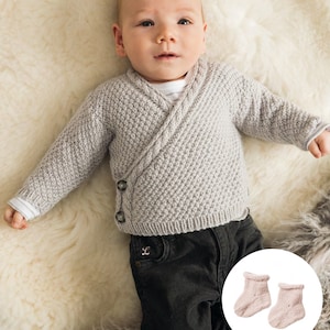 Peut inclure: Un bébé portant un cardigan gris en tricot avec un devant croisé et un pantalon noir. Le bébé est allongé sur une surface blanche et moelleuse. Une paire de chaussons en tricot rose pour bébé est représentée dans un cercle sur le côté droit de l'image.