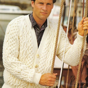 Op de afbeelding: Een witte kabelgebreide cardigan met een knoopsluiting en lange mouwen. De cardigan is gemaakt van dik garen en heeft een textuurpatroon.