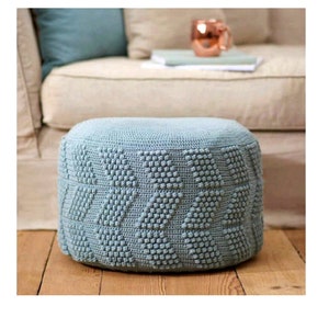 May include: A light blue crocheted footstool with a textured diamond pattern. The footstool is sitting on a wooden floor.
