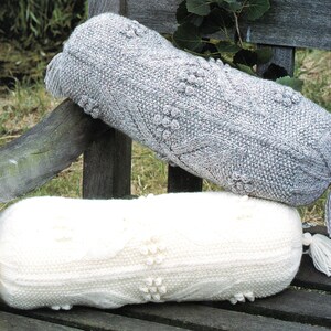 May include: Two knitted bolster pillows with tassels. The top pillow is gray and the bottom pillow is white. Both pillows have a textured pattern.