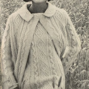 May include: A woman wearing a white cable knit sweater with a collar. She is standing in a field of tall grass.