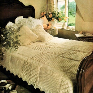 May include: A white crocheted bedspread with a delicate scalloped edge, draped over a wooden bed frame with a white ruffled headboard. The bed is decorated with white pillows and a bouquet of flowers in a vase on a side table.