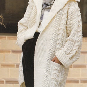 以下が含まれることがあります： クリーム色の長めのニットカーディガン。ショールカラーとポケット付き。 ケーブル編みとポコポコとしたデザインが特徴です。 袖口はリブ編み。白いトップスと黒いパンツの上に着用されています。