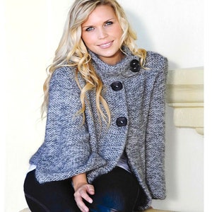 May include: A woman wearing a grey and white knitted sweater with black buttons and black trousers. She is sitting on a white surface.