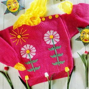 May include: A pink knitted sweater with a white daisy and green stem design. The sweater has yellow smiley face buttons and a yellow sun with a smiley face in the center.
