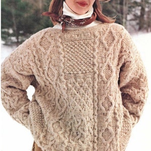 Może przedstawiać: Beżowy sweter z warkoczami, brązowym kapeluszem i okularami przeciwsłonecznymi. Sweter ma fakturowany wzór i luźny krój.