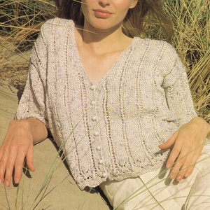 May include: A white knitted cardigan with a v-neck and pearl buttons. The cardigan has a delicate openwork pattern and is being worn by a person with long hair.