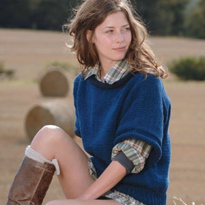 Puede incluir: Una mujer con un suéter de punto azul y una camisa a cuadros debajo, sentada en una paca de heno en un campo.