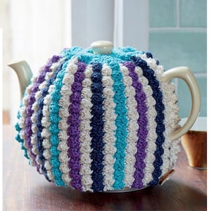 Puede incluir: Un cubre-tetera de ganchillo en tonos turquesa, morado, blanco y azul marino. El cubre-tetera está hecho con un punto texturizado que crea una superficie abultada.