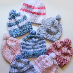 May include: Eight knitted baby hats in various colors and patterns, including pink, blue, and white stripes. The hats have pom-poms on top.