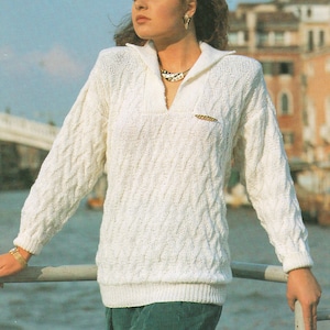 May include: A white knitted jumper with a cable knit pattern and a collared neckline. The jumper has a gold button closure at the neckline.