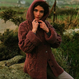 May include: A maroon knitted cardigan with a hood and button fastening. The cardigan features a cable knit design and is being modelled by a person.