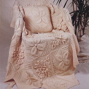 May include: A cream-colored knitted blanket and pillow set draped over a chair. The blanket features large, raised floral designs and tassels. The pillow has a central flower motif. The set is made of a soft, textured yarn.