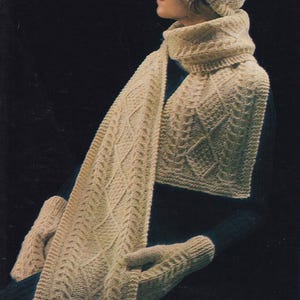 May include: A cream-colored knitted hat, scarf, and mittens set. The hat has a pom-pom on top. The scarf is long and has a cable knit pattern. The mittens are fingerless.