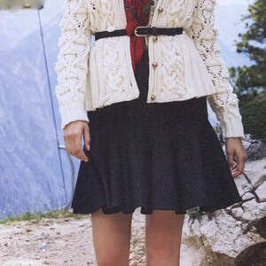 May include: A woman wearing a white cable knit cardigan with a red floral scarf, a black belt, and a black skirt. She is wearing black hiking boots with gold accents.