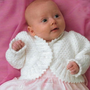 May include: A white knitted baby cardigan with a scalloped edge and button closure. The cardigan is worn by a baby lying on a pink blanket.