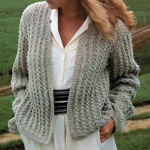 Könnte beinhalten: Ein hellgrauer Cardigan mit offenem Vorderteil und einem zarten Strickmuster. Der Cardigan wird über einem weißen Oberteil und einem schwarz-weiß gestreiften Gürtel getragen.