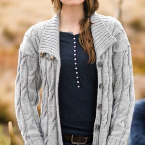 May include: A grey cable knit cardigan with a large collar, long sleeves, and a button fastening. The cardigan has pockets on the front and is worn over a black t-shirt and blue jeans.