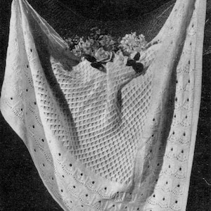 Pode incluir: Um cobertor de bebé branco de crochê com um delicado padrão de renda. O cobertor está drapeado sobre uma cesta de flores.