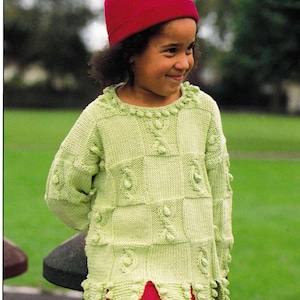 May include: A young girl wearing a green knitted sweater with a checkered pattern and a red beanie. The sweater has a scalloped hem and a textured design.