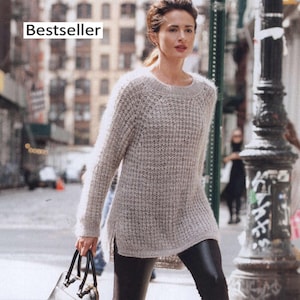 Puede incluir: Un suéter de punto gris claro con un patrón texturizado y mangas largas. El suéter tiene un cuello redondo y un dobladillo con abertura. La palabra "Bestseller" está en un cuadro blanco en la esquina superior izquierda. La persona lleva leggings negros y un bolso negro.