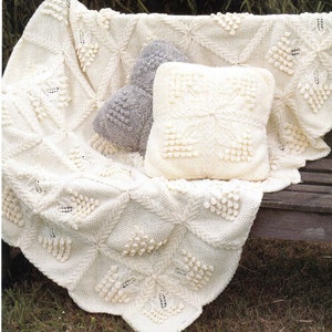 May include: A white crocheted throw blanket with a textured pattern and a white crocheted pillow with a similar pattern. The blanket and pillow are sitting on a wooden bench.