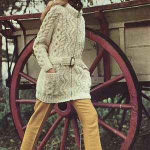 May include: A woman wearing a white cable knit cardigan with a brown belt and a brown and white polka dot headscarf. She is standing next to a red wooden wagon wheel.