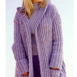 May include: A lavender knitted cardigan with a hood and long sleeves, featuring two front pockets. The cardigan is worn over a lavender turtleneck and trousers. The knitwear has a ribbed texture.