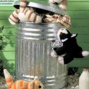 May include: Three knitted cat toys in a variety of colors and patterns, including tabby, black and white, and orange and white. The toys are posed around a metal trash can and a glass milk bottle.