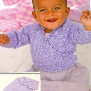 May include: A baby wearing a light purple knitted wrap sweater and a matching skirt. The sweater has a wrap design with two white buttons. The skirt has a decorative lace trim. The baby is smiling.