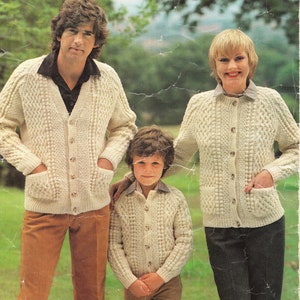 Può includere: Una famiglia di tre persone che indossano cardigan di lana color crema con chiusura a bottoni. I cardigan hanno un motivo a treccia e tasche. La famiglia è in piedi in un campo erboso.