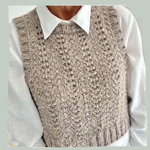 May include: A beige knitted vest with a textured pattern. The vest is worn over a white collared shirt. The knitted vest has a ribbed hem and a detailed, vertical design. The overall style is casual and comfortable.