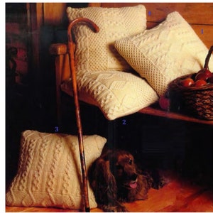 以下が含まれることがあります： ケーブル編みのパターンが施されたクリーム色のニット製のクッションが4つあります。2つのクッションは木製のベンチの上にあり、1つは床にあり、もう1つは木製のテーブルの下にあります。茶色の木製の杖がベンチに寄りかかっています。茶色の犬がクッションの前で床に横たわっています。