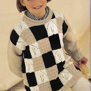 May include: A child wearing a beige, black, and white checkered sweater with a lace pattern. The sweater has a round neckline and long sleeves.