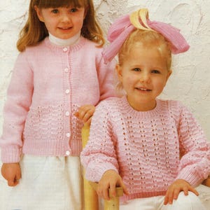 May include: Two young girls wearing pink knitted cardigans. One cardigan has a button-up front and the other is a pullover style. Both cardigans feature a delicate lace pattern.