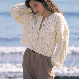 May include: A cream-coloured, cable-knit cardigan with a button-up front and long sleeves. The sweater has a textured pattern and is worn with brown trousers. The background is a blurred ocean scene.