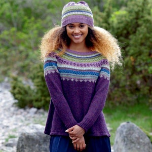 Könnte beinhalten: Eine Frau trägt einen lila Pullover mit einem bunten gemusterten Joch und eine passende lila und grüne Strickmütze. Sie steht vor einem grünen Hintergrund.