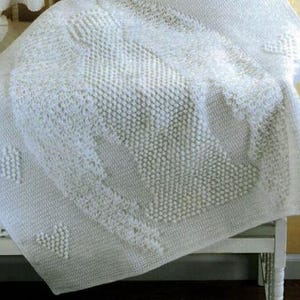 May include: A white crocheted blanket with a textured design featuring a heart pattern. The blanket is folded and draped over a white chair.