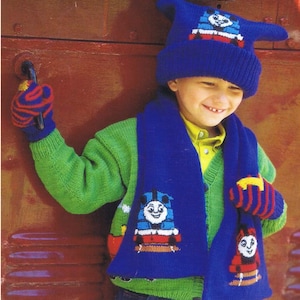 May include: A blue knitted scarf with a Thomas the Tank Engine design. The scarf is worn by a child wearing a matching blue knitted hat with a Thomas the Tank Engine design. The child is also wearing a green sweater and red and blue striped mittens.