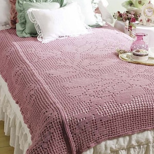 May include: A crocheted throw blanket in a dusty rose color with a delicate open stitch pattern. The blanket is draped over a bed with white ruffled bedding.