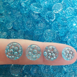 May include: A collection of small, round, translucent light blue glass cabochons featuring a textured surface with raised bubble-like patterns. Four individual cabochons are displayed in a row, showing varying sizes of the same circular design.