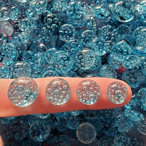 May include: Four clear, round, blue glass beads with internal bubbles are held on a finger. The beads are surrounded by a larger collection of similar beads, creating a textured, aquatic appearance. The beads are likely decorative.
