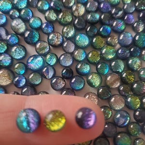 Può includere: Una collezione di perline di vetro iridescente in tonalità di blu, verde e viola. Le perline sono rotonde e hanno una superficie leggermente strutturata. Tre perline sono evidenziate su un dito, mostrando una perlina verde, blu e viola.