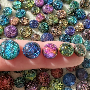 May include: A collection of iridescent, glittery glass marbles in various colors, including blue, green, purple, and gold. Some marbles are held on a finger, showcasing their round shape and sparkling texture. The marbles are scattered on a white surface.